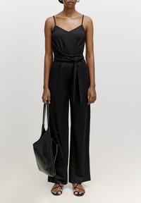 Schwarzer Jumpsuit mit Spaghettiträgern, tailliertem Bund, weiter Beinform, kombiniert mit flachen schwarzen Sandalen und einer schwarzen Tote-Bag. Glattes Material.