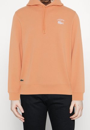 Osoba w morelowej bluzie z kapturem Lacoste z logo na piersi i małym emblematem krokodyla przy dole, połączona z czarnymi spodniami.