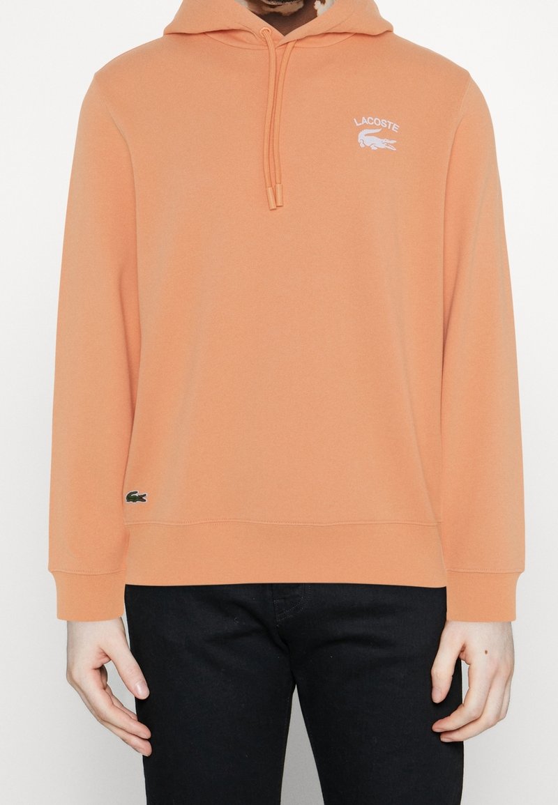 Personne portant un sweat à capuche Lacoste pêche avec logo sur la poitrine et petit emblème crocodile près de l'ourlet, associé à un pantalon noir.