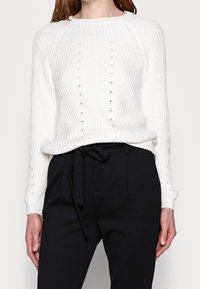 Witte gebreide trui met raglanmouwen, voorzien van verticale uitsparingen en ribgebreide manchetten. Gecombineerd met zwarte high-waisted broek en een gestrikt tailleband.