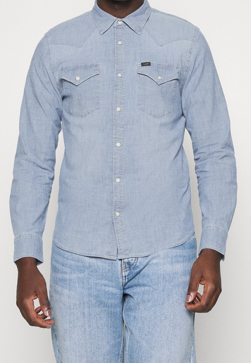Chemise en denim bleu clair à manches longues, col boutonné, deux poches poitrine et boutons-pression. Présente une texture légèrement délavée.