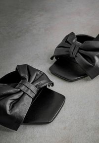 Sandales en cuir noir avec un design à bout carré, présentant un accent de nœud proéminent sur le dessus. Texture lisse avec une finition mate.