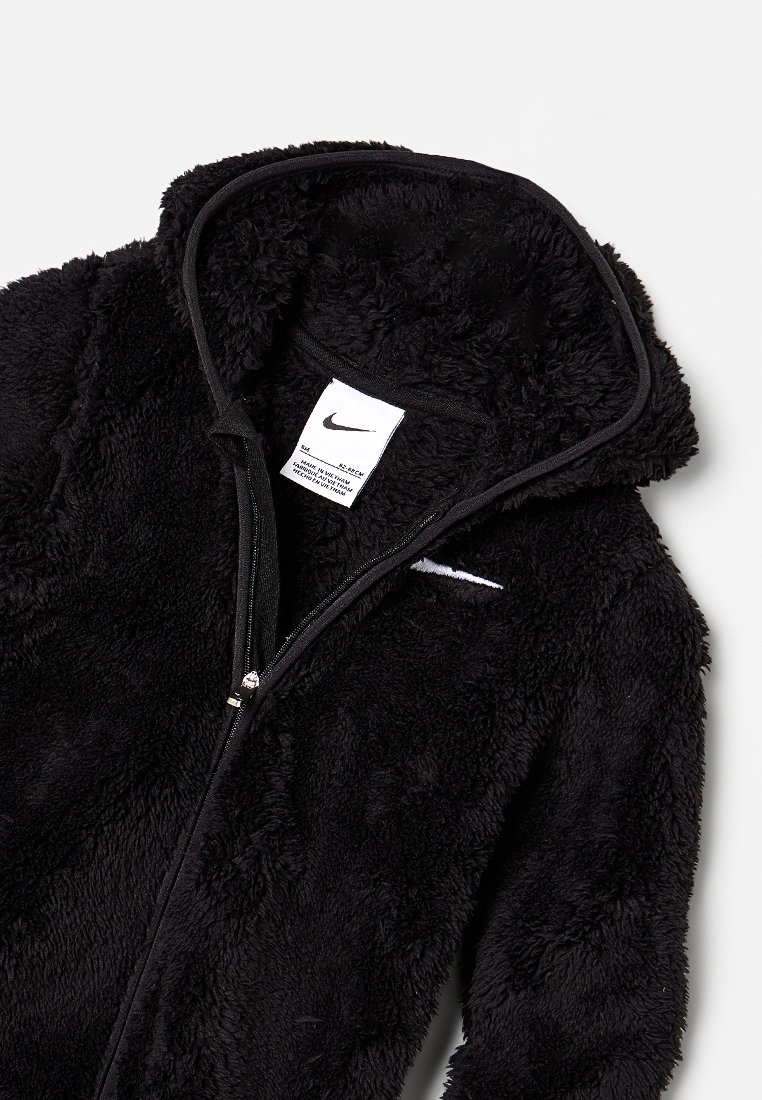 Chaqueta de forro polar negra con cremallera y capucha, con un tejido suave y texturizado y un pequeño logo blanco de Nike en el pecho. Etiqueta de cuidado visible en el interior.