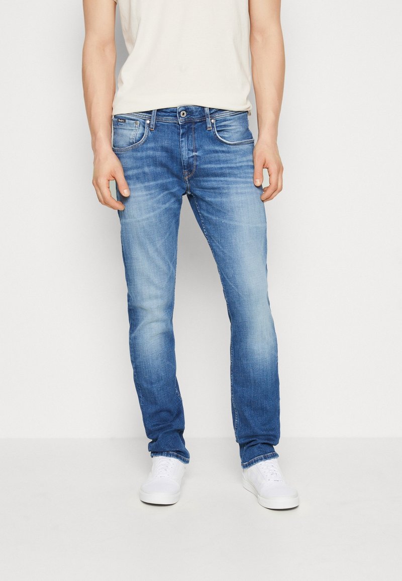 Vaqueros de mezclilla azul con un acabado desgastado, diseño de pierna recta, cinco bolsillos y detalles de costura en contraste. Combinados con zapatillas blancas.