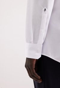 Witte lange mouwen shirt met een knoopmanchet. Bevat een zwart geborduurd roosdetail op de manchet. Gladde, lichte stof met een getailleerde pasvorm.