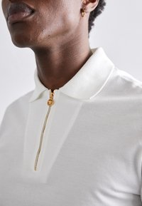 Balta polo krekla ar zelta rāvējslēdzēju, apkakli un teksturētu audumu. Tuvplānā izceltas šuvju un rāvējslēdzēja detaļas.