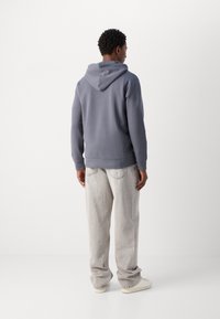 Siva kapuca s kenguresko žepom i kapuljačom na vezanje, u kombinaciji sa svijetlosivim ravnim trapericama, prikazano s leđa. Teksturirana tkanina.