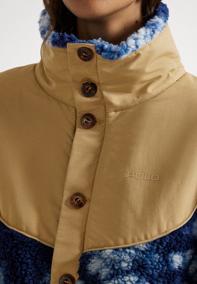Μπεζ τζάκετ με γαλάζιο κολάρο από φλις. Διαθέτει πέντε καφέ κουμπιά και το λογότυπο "POLO" χαραγμένο στο ύφασμα. Μαλακή υφή και στρωματοποιημένος σχεδιασμός.