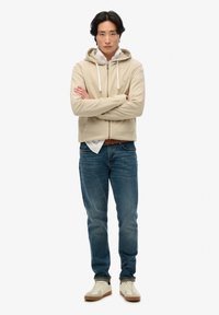 Sudadera beige con cremallera y cordones blancos, combinada con una camiseta blanca. Llevada con vaqueros azules y zapatillas color crema con detalles marrones.