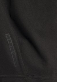 Sort stof med en glat tekstur har en lodret gummietiket, der står "UANSET TESTEN" i præget skrifttype.