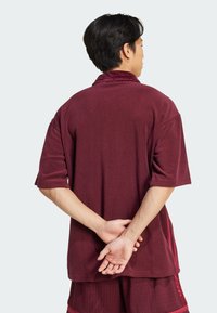 Camisa corta de manga corta color burdeos hecha de una tela suave, con un corte holgado y un cuello doblado. Usada con las manos entrelazadas detrás.