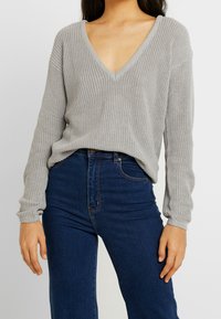 Pull en tricot gris clair avec un décolleté en V profond et des manches longues, associé à un jean taille haute bleu foncé. Tissu texturé et coupe décontractée.