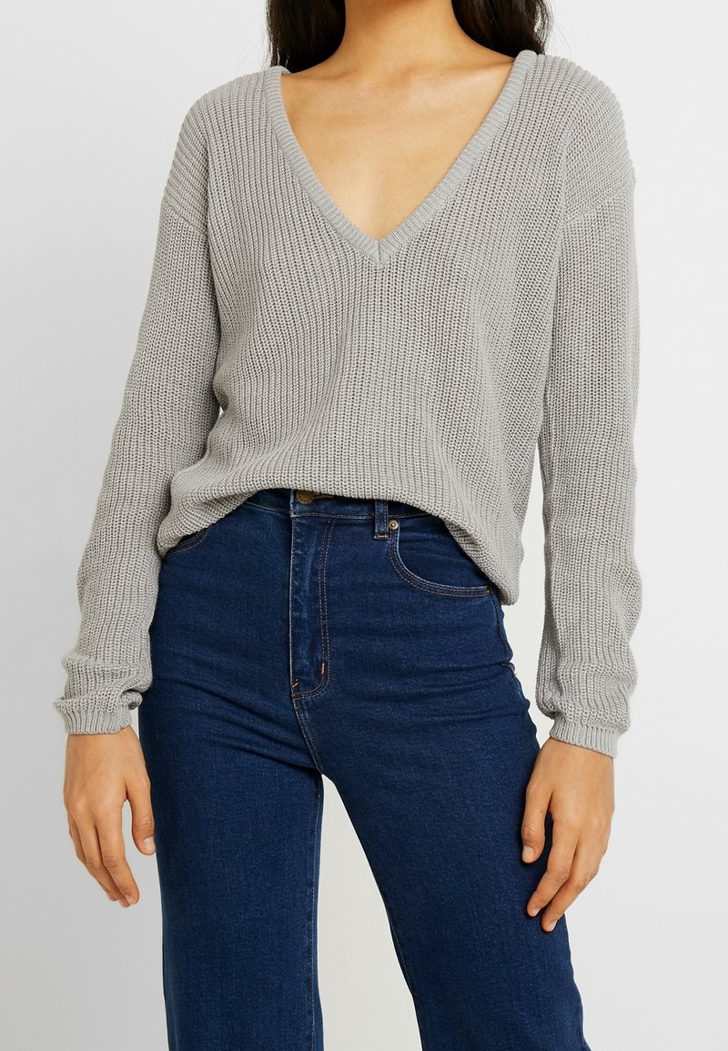 Pull en tricot gris clair avec un décolleté en V profond et des manches longues, associé à un jean taille haute bleu foncé. Tissu texturé et coupe décontractée.