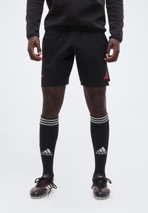 Mann steht in schwarzen Adidas Fußballshorts, schwarzen Adidas knielangen Socken mit weißen Streifen und schwarzen Adidas Fußballschuhen mit roten Akzenten.