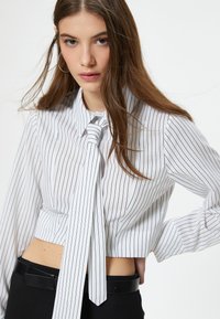 Camicia bianca a maniche lunghe con strisce verticali nere, dotata di un nodo al collo. Indossata con una cintura nera. Tessuto liscio.