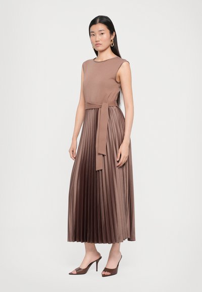 Long, pleated dress in brown fabric, featuring a fitted bodice and a tied waist. The model wears open-toe high heels.