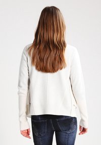 Krämfärgad stickad cardigan med långa ärmar, avslappnad passform, ribbade muddarna och sidofickor, kombinerat med mörka jeans.