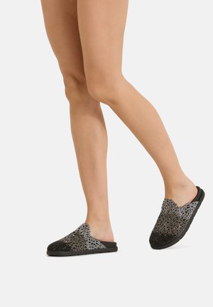 Gambe che indossano scarpe slip-on nere con intricati motivi di strass argento e bordi smerlati, camminano su uno sfondo chiaro e uniforme.