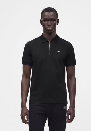 Homme portant un polo noir à manches courtes avec une fermeture éclair argentée et un petit logo sur la poitrine, debout devant un fond clair uni.