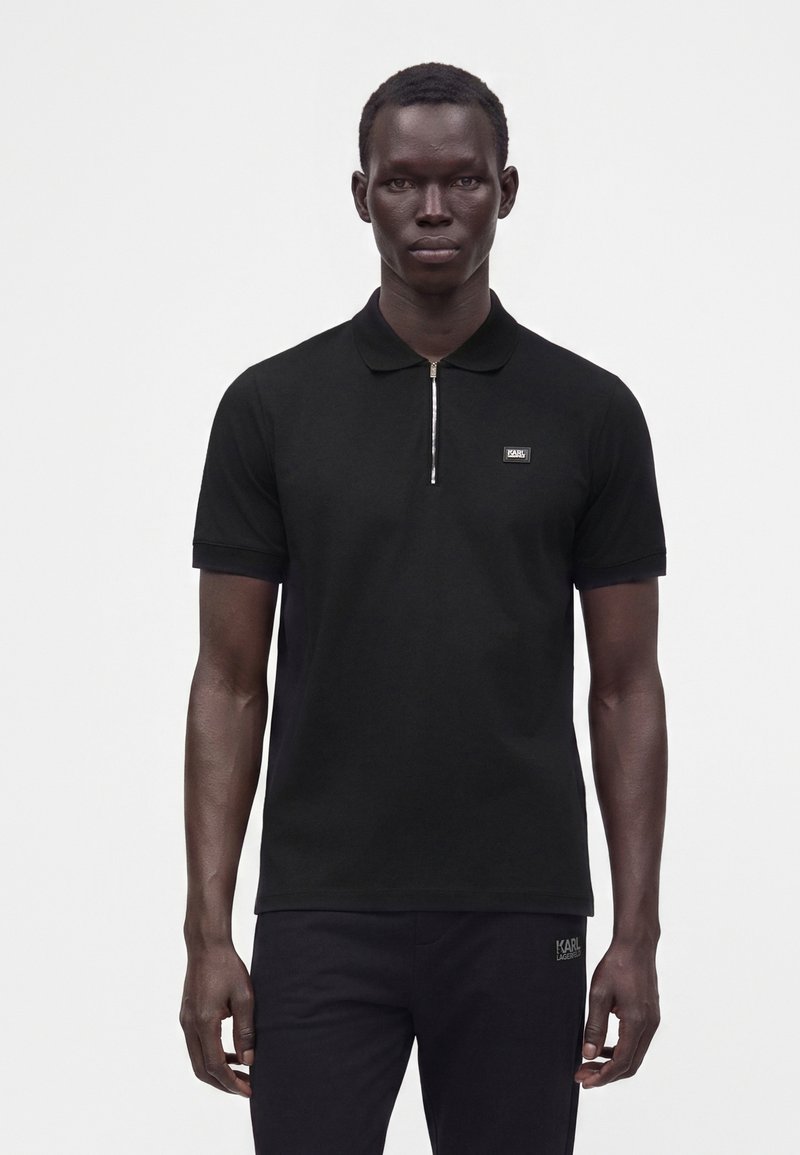 Homme portant un polo noir à manches courtes avec une fermeture éclair argentée et un petit logo sur la poitrine, debout devant un fond clair uni.