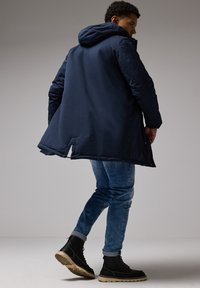 Chaqueta con capucha de color azul marino y corte largo; presenta una abertura en la parte trasera. Lleva puesta pantalones vaqueros azul claro y botas negras con suela robusta.