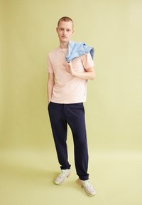 Lichtroze t-shirt, marineblauwe broek en multicolore sportschoenen. Spijkerjack over de schouder gedrapeerd. Eenvoudige, casual fit tegen een zachte groene achtergrond.