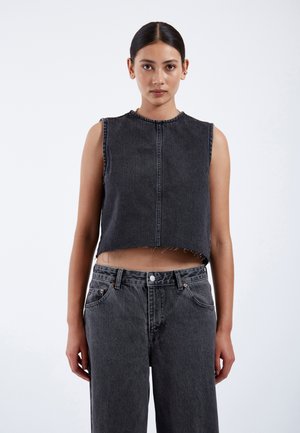 Frau mit dunklen Haaren in einem tiefen Dutt, die ein ärmelloses schwarzes kurzes Denim-Top und hoch taillierte schwarze Denim-Jeans trägt, stehend vor einem schlichten weißen Hintergrund.