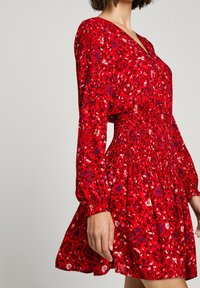 Robe fleurie rouge avec manches longues, taille smockée et jupe évasée. Présente des motifs de fleurs roses et bleues sur un tissu texturé.