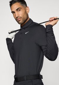 Polo noir manches longues avec une légère brillance, doté d'un col et d'un logo Nike blanc. Porté avec une ceinture noire, tenant un club de golf.