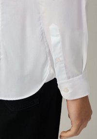 Witte lange mouwen shirt met een glad, geweven textuur, knoopmanchetten en een afgeronde zoom. Op een zwarte achtergrond is de vorm van het shirt zichtbaar.