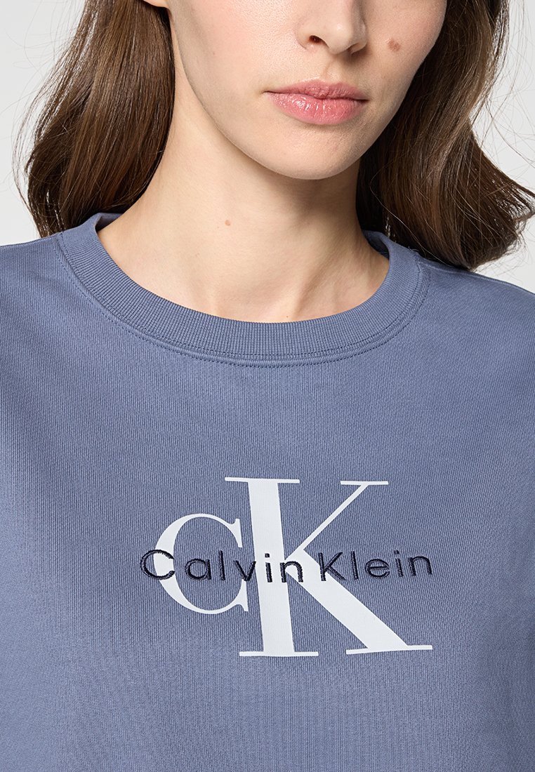 Felpa in cotone azzurro chiaro con colletto a girocollo e un grande logo bianco con la scritta "CK" e "Calvin Klein". Texture morbida, vestibilità casual.