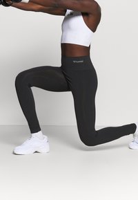 Osoba w czarnych legginsach i białej bluzce krótki z przodu wykonuje krok w przód z wyciągniętymi ramionami, mając na sobie białe sneakersy i białe skarpetki na jednolitym tle.