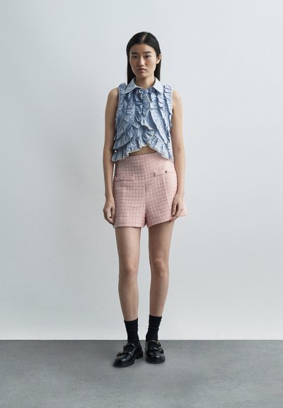 Γυναίκα που φοράει μπλε μπλούζα χωρίς μανίκια με βολάν, ροζ σορτς με υφή, μαύρες κάλτσες και μαύρα μοκασίνια, στέκεται πάνω σε γκρι πάτωμα με απλό ανοιχτό φόντο.