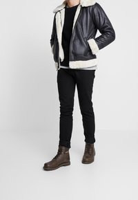 Czarna skórzana kurtka z kremowym futerkiem podszytym i mankietami, w połączeniu z czarnym swetrem, czarnymi spodniami i brązowymi butami na sznurówki.