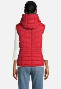 Vrouw met donker haar in een bun, gekleed in een rode gewatteerde hoodie-vest over een witte lange mouw top en blauwe spijkerbroek, met haar rug naar de kijker toe.
