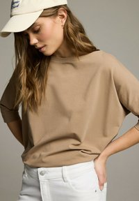 Jasnobrązowa, luźna koszula z rękawami do trzech czwartych, w połączeniu z białymi spodniami. Krema kolorowa czapka z niebieskim haftem uzupełnia całość.