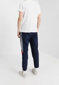 Pantalones de chándal navy con rayas blancas, ajuste ceñido, puños elásticos y acentos rojos en los lados. Combinados con una camiseta blanca sencilla y zapatos claros.