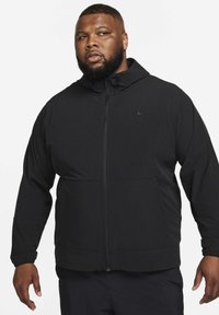 Zwarte, gevoerde jas gemaakt van lichtgewicht materiaal, met een ritssluiting aan de voorkant en elastische manchetten. Voorzien van een subtiel Nike-logo op de linkerborst.