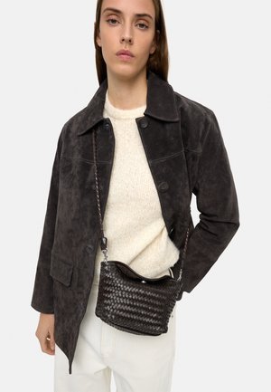 Kobieta w ciemnej zamszowej kurtce, kremowym dzianinowym swetrze, białych spodniach, niosąca małą czarną plecioną torebkę na ramię z plecionym paskiem.