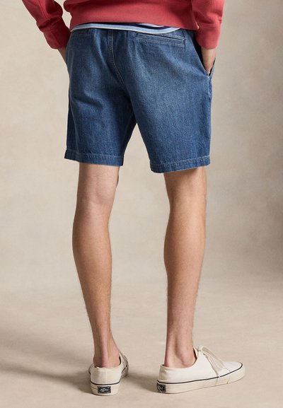 Άνδρας στέκεται με τα χέρια στις τσέπες, φορώντας μπλε τζιν σορτς, κόκκινο φούτερ και λευκά πάνινα παπούτσια σε ουδέτερο φόντο.