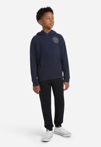 Μπλε ναυτικό hoodie με τσέπη μπροστά και λεπτομέρεια λογότυπου, συνδυασμένο με μαύρα παντελόνια και άσπρα αθλητικά παπούτσια. Μαλακό υλικό και άνετη εφαρμογή.