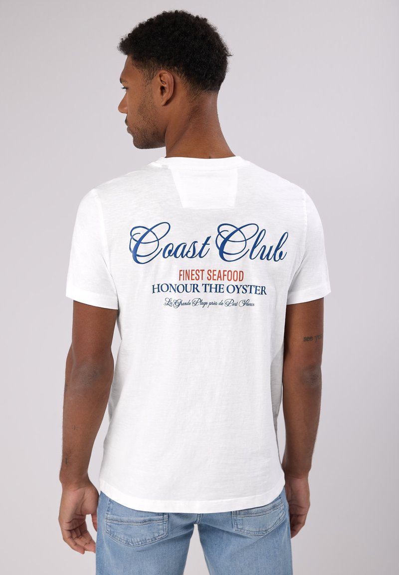 Bílý bavlněný tričko s krátkým rukávem, na zadní straně s nápisem "Coast Club" v modrém a červeném písmu, uvolněný střih a minimální textura.
