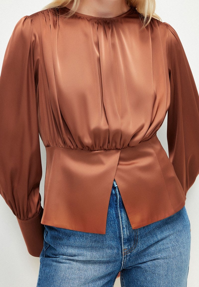 Blouse en satin marron avec manches bouffantes, froncée à la taille et fente à l'avant. Idéale pour un duo avec du denim.