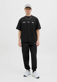 Camiseta negra de gran tamaño con gráficos blancos y un logo negro, combinada con pantalones jogger negros. El modelo lleva una gorra gris y zapatillas blancas.