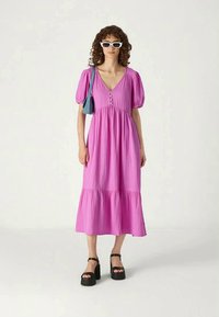 Robe rose, midi avec des manches bouffantes, un décolleté en V et des boutons décoratifs. Présente un design à volants et est associée à des sandales compensées.