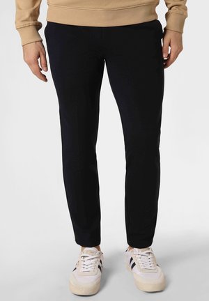 Persoon die zwarte slim-fit broek, beige trui en witte sneakers met zwarte strepen draagt, staand tegen een effen lichte achtergrond.