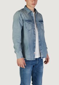 Camicia in denim azzurro chiaro con design a bottoni, due tasche sul petto e colletto classico. Indossata sopra una maglietta bianca, con maniche lunghe.