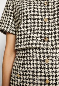 Personne portant une veste à manches courtes et une jupe à motif pied-de-poule noir et blanc avec des boutons dorés, vue de l'épaule à la taille.