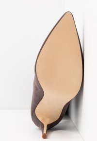 Scarpa con tacco alto in suede marrone a punta, poggiata sul lato che mostra la suola liscia color beige e la sottile punta del tacco a stiletto.