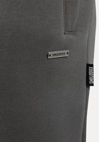 Tejido atlético gris con textura acanalada. Presenta una etiqueta de metal con "SMILODOX" y una etiqueta negra en el costado. Diseño ajustado, estilo minimalista.